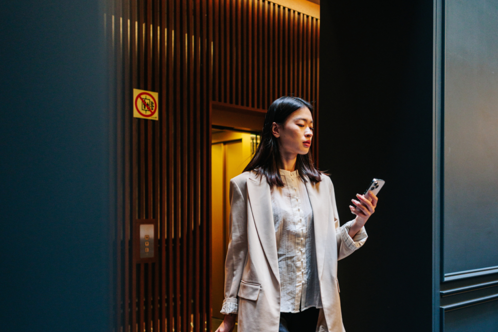 Woman walking through a hotel space with a suitcase while checking her phone.